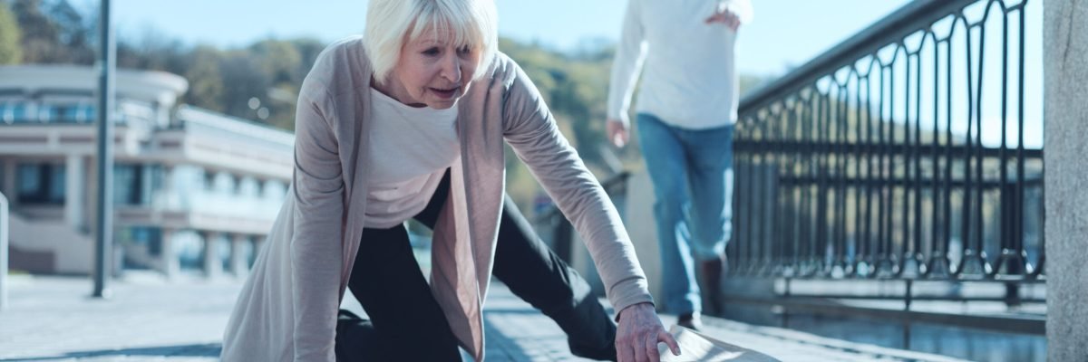 Are you ok. Selective focus on a scared senior lady standing on her knee and trying to pick up her groceries after falling down while her worried husband running to her in the background.