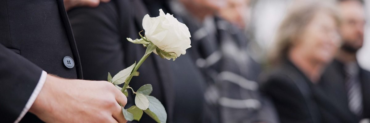 Hands, rose and a person at a funeral in a cemetery in grief while mourning loss at a memorial service. Death, flower and an adult in a suit at a graveyard in a crowd for an outdoor burial closeup